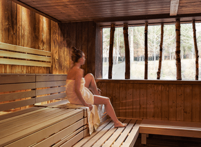Eine Frau sitzt in einer holzverkleideten Damen Sauna, blickt durch ein Fenster mit natürlichem Licht und Aussicht auf Bauume. Sie ist in ein Handtuch gewickelt und entspannt sich in der ruhigen Atmosphaere.