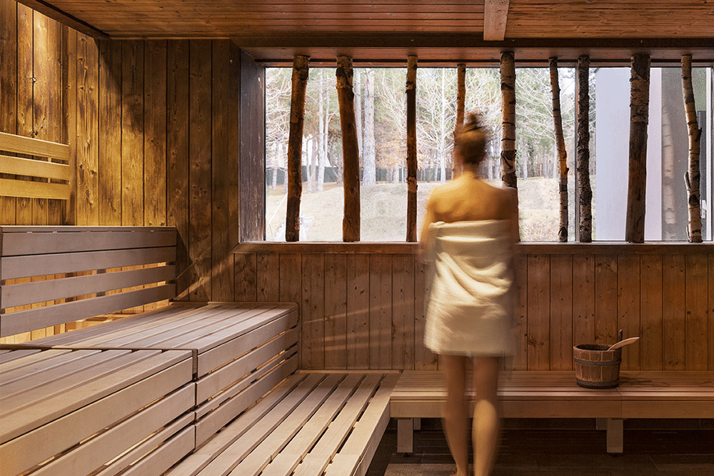 Junge Frau mit Handtuch um den Koerper steht mit dem Ruecken in die Kamera in einer Sauna der Therme Wien und blickt aus dem Fenster der Sauna.