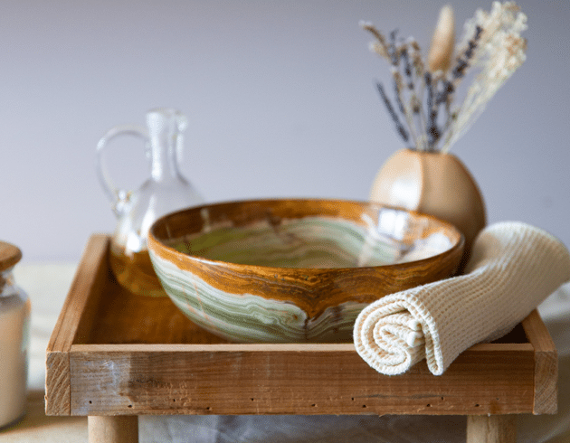 Ein entspannendes Wellness-Set-up auf einem Holztisch in der Therme Wien, bestehend aus einer gemusterten Keramikschale, einer durchsichtigen Karaffe mit Oel, einem gefalteten weissen Handtuch und getrockneten Lavendelblueten in einer Vase.
