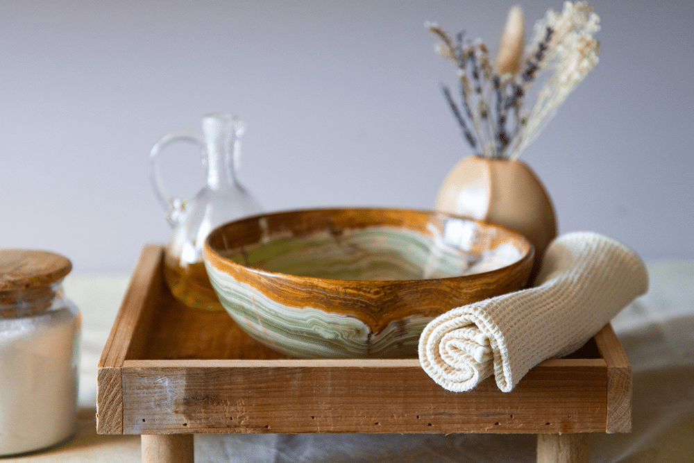 Ein entspannendes Wellness-Set-up auf einem Holztisch in der Therme Wien, bestehend aus einer gemusterten Keramikschale, einer durchsichtigen Karaffe mit Oel, einem gefalteten weissen Handtuch und getrockneten Lavendelblueten in einer Vase.