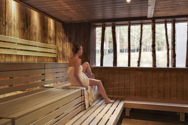 Junge Frau mit Handtuch um den Koerper sitzt in einer Sauna der Therme Wien und blickt aus dem Fenster der Sauna.